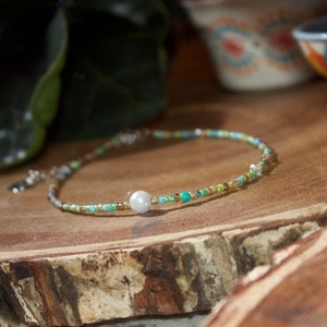 May include: A delicate beaded necklace with a single pearl accent. The necklace is made with small, colorful beads in shades of green, blue, and gold. The pearl is white and sits in the center of the necklace.