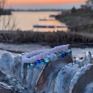 May include: Two bracelets with white and blue beads. The bracelets are on a silver-colored surface. The background is a blurry sunset over a lake.