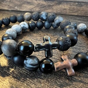 May include: Two beaded bracelets with cross charms. One bracelet features black onyx, lava rock, and gray stone beads, with a black cross charm. The other bracelet has similar beads and a copper cross charm.