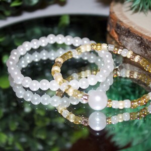 May include: Three white and yellow beaded bracelets with a large white pearl charm. The bracelets are stacked on top of each other.