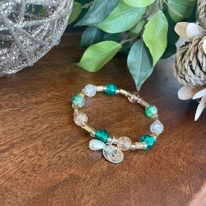 May include: A green and gold beaded bracelet with a silver charm featuring a shamrock. The bracelet is made with small, round beads and has a delicate, feminine look.