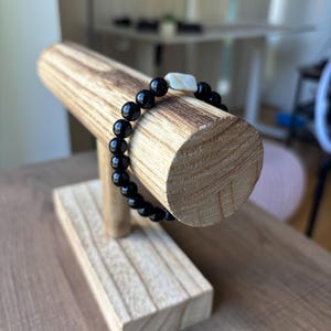 May include: A black beaded bracelet with a white rectangular accent bead is displayed on a wooden jewelry stand. The bracelet is made of round, glossy black beads. The stand is made of light-colored wood.