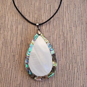 Op de afbeelding: Een traanvormige hangerketting met een wit parelmoer centrum en een rand van iriserende abalone schelp. De ketting is aan een zwart lederen koord.