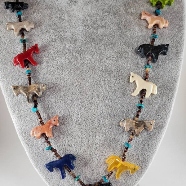 May include: A necklace made of carved stone horses in various colors, including red, brown, black, white, and yellow. The horses are strung on a brown cord with turquoise beads.