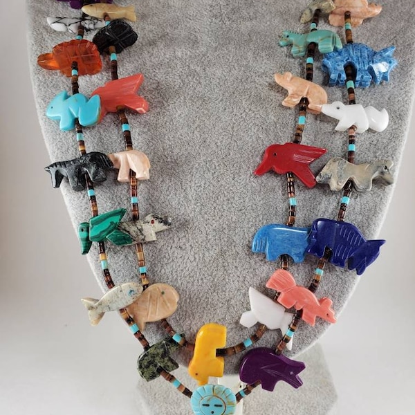 May include: A multi-strand necklace featuring carved animal figurines made from various stones, including turquoise, coral, and jasper. The necklace is strung with brown beads and features a small carved figurine of a person at the bottom.