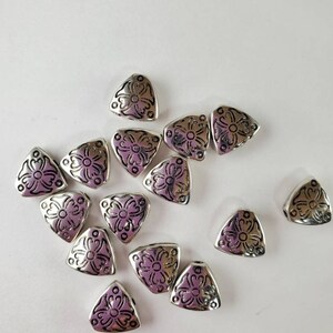 May include: A collection of silver triangle-shaped beads with a floral design. The beads are scattered on a white surface.