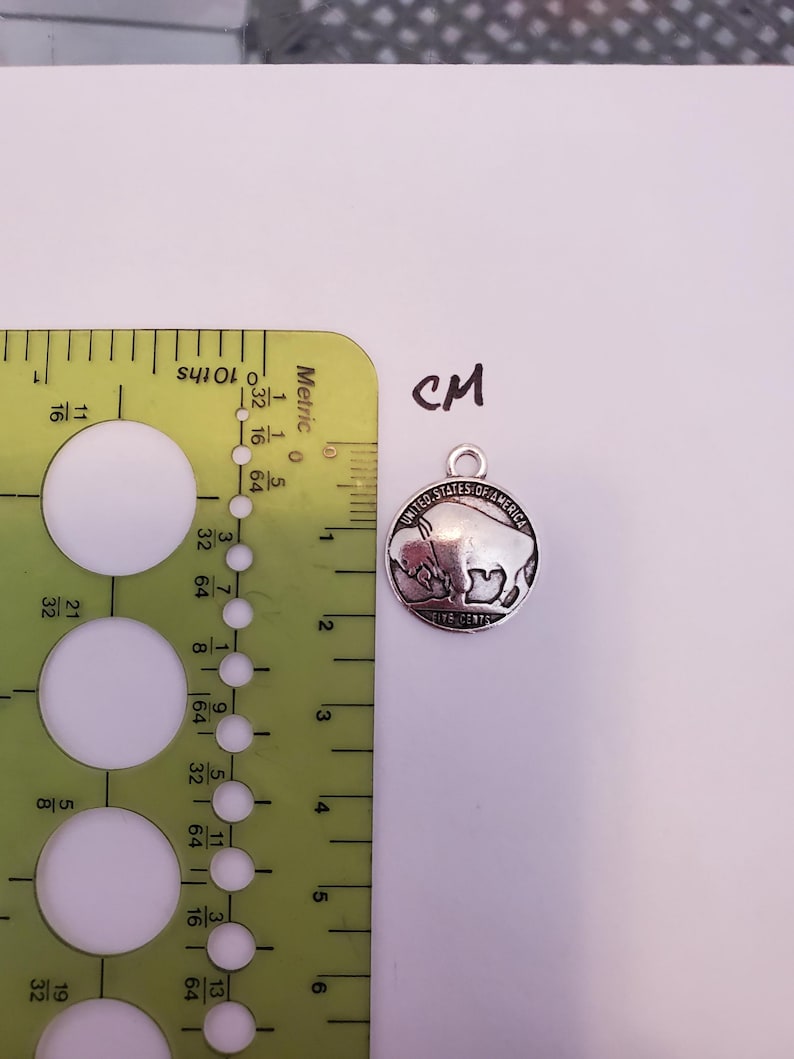 May include: A silver-toned charm featuring a buffalo silhouette with the text "United States of America Five Cents" around the edge. The charm is approximately 2 cm in diameter.