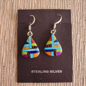 May include: A pair of sterling silver earrings with a teardrop-shaped design. The earrings are made with colorful inlay stones, including turquoise, coral, and lapis lazuli. The earrings are on a black card with the text "STERLING SILVER".