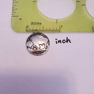 May include: A silver-colored United States five-cent coin with the inscription "United States of America" and "Five Cents" on the back. The coin is placed on a white surface next to a ruler with markings in inches.