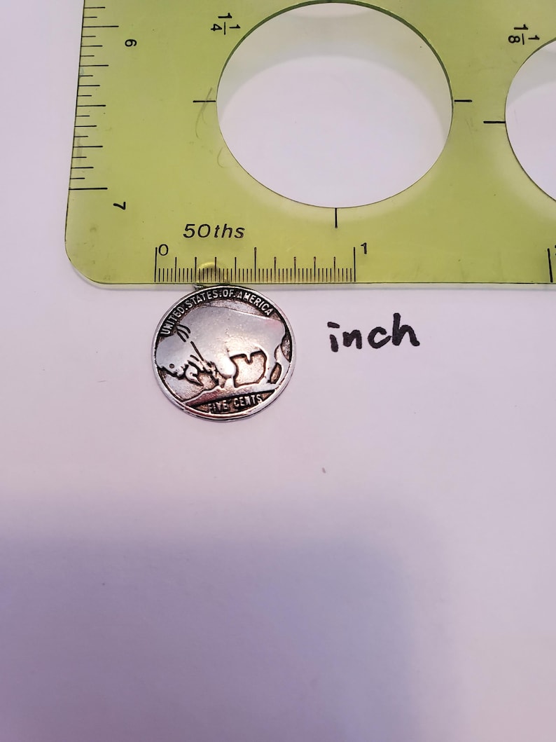 May include: A silver-colored United States five-cent coin with the inscription "United States of America" and "Five Cents" on the back. The coin is placed on a white surface next to a ruler with markings in inches.