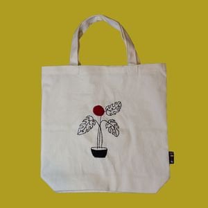 Könnte beinhalten: Eine weiße Stofftasche mit einem schwarzen gestickten Design einer Topfpflanze mit drei Blättern und einer roten Blüte.