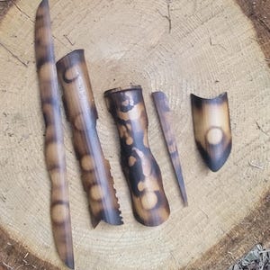 May include: A collection of handcrafted bamboo pieces with a dark, burnt finish, arranged on a cross-section of a tree trunk. The pieces vary in shape and size, showcasing natural wood grain and textures. The background includes foliage.