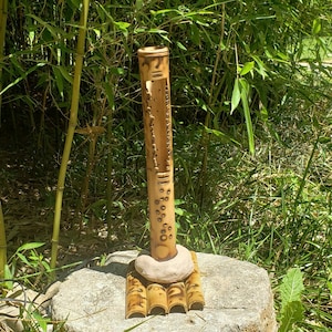 May include: A bamboo incense burner with a carved design and a round stone base. The burner is sitting on a stone platform surrounded by green grass and brown leaves.