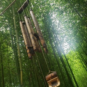 May include: A hanging wind chime constructed from multiple hollow bamboo tubes of varying lengths, featuring natural brown and tan wood grain patterns, suspended by thin cords with a curved bamboo striker and a decorative bamboo wind catcher.