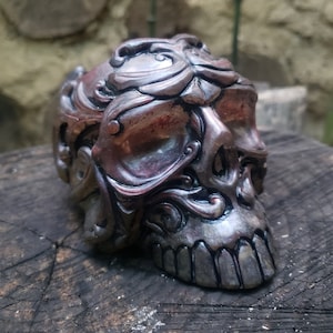 May include: A detailed, ornate skull sculpture with intricate carvings and a metallic finish. The skull is painted in shades of silver, black, and red, giving it a gothic and decorative look.
