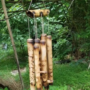 May include: A hanging wind chime constructed from multiple hollow bamboo tubes of varying lengths. The bamboo features natural tan and brown coloration with dark charred markings and etched geometric patterns, suspended by white strings from a top bar.