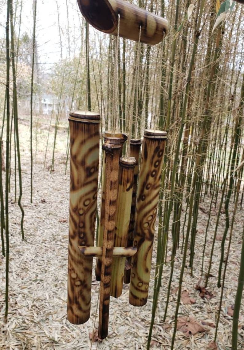 May include: A bamboo wind chime featuring six hollow cylindrical tubes of varying lengths suspended from a horizontal bamboo piece by thin cords. The tubes are light tan with dark brown scorched patterns, including circles and wavy lines.