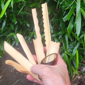May include: A collection of five light tan bamboo tools with varied carved edges, including straight, wavy, and serrated designs, held together in a dark brown bamboo cylindrical base with a smooth, polished finish and visible wood grain.