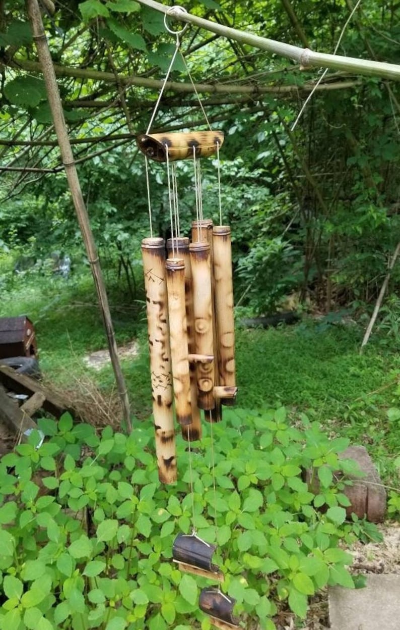 May include: A hanging wind chime constructed from multiple hollow bamboo tubes of varying lengths. The bamboo features natural tan and brown coloration with dark charred markings and etched geometric patterns, suspended by white strings from a top bar.