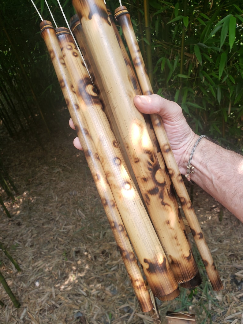 May include: A wind chime constructed from multiple cylindrical bamboo tubes of varying lengths. Each tube features a natural light tan finish with dark brown, scorched circular markings and patterns. The tubes are suspended by thin white cords.