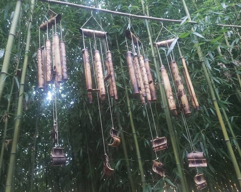 May include: Four bamboo wind chimes hanging from a horizontal wooden rod. Each chime consists of multiple hollow bamboo tubes of varying lengths, featuring dark brown etched patterns and circular designs, suspended by thin white strings.
