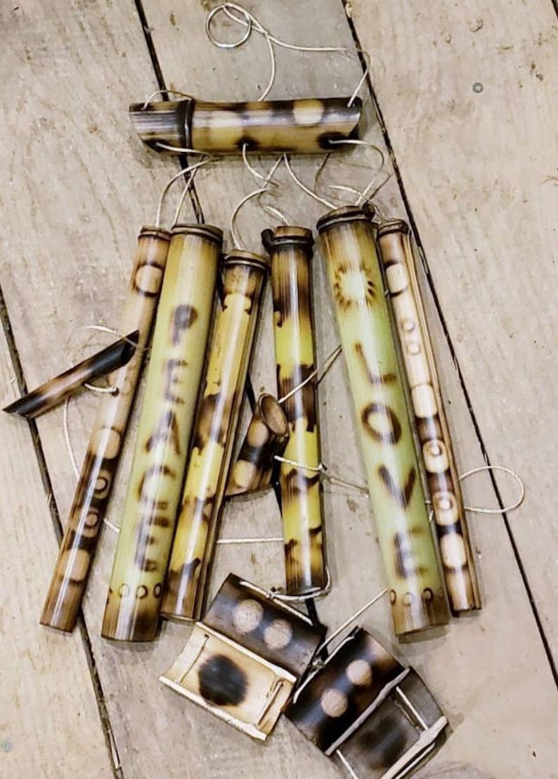May include: A bamboo wind chime featuring multiple hollow tubes suspended by strings from a horizontal bamboo bar. The tubes are decorated with dark brown mottled patterns and the words PEACE and LOVE burned into the light tan bamboo surface.