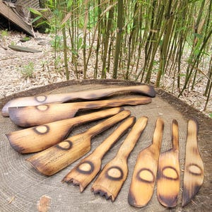 May include: A collection of handmade wooden kitchen utensils, including spatulas and stirring spoons, with a burnt design. The utensils are arranged on a tree stump, with bamboo stalks in the background.