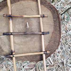 May include: A small, handcrafted bamboo ladder with four rungs, connected with black twine. The ladder rests on a large, circular wooden slice. The bamboo has a natural, light brown color with darker, burnt accents.