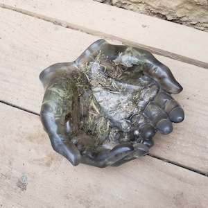 May include: A gray resin hand sculpture with a textured, marbled finish. The hand is cupped, forming a bowl shape.