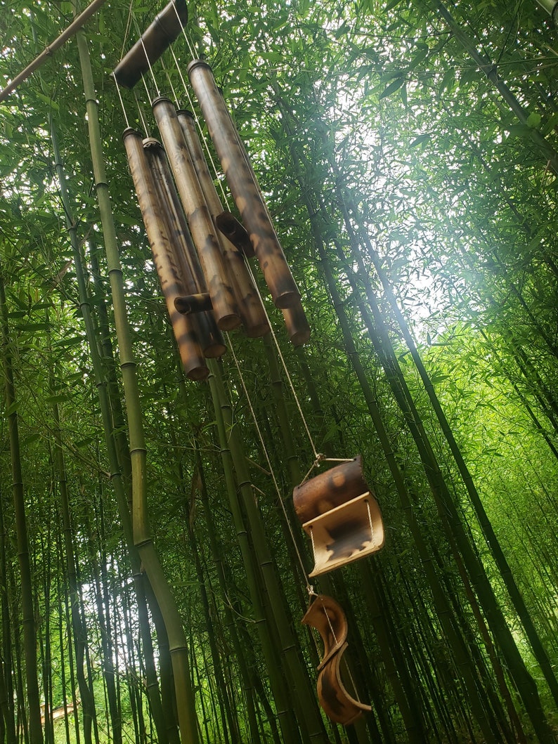 May include: A hanging wind chime constructed from multiple hollow bamboo tubes of varying lengths, featuring natural brown and tan wood grain patterns, suspended by thin cords with a curved bamboo striker and a decorative bamboo wind catcher.