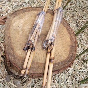May include: Two bundles of bamboo poles, each wrapped in clear plastic. The bamboo has a natural, light brown color with darker, charred sections at the ends. The poles are resting on a wooden tree stump.