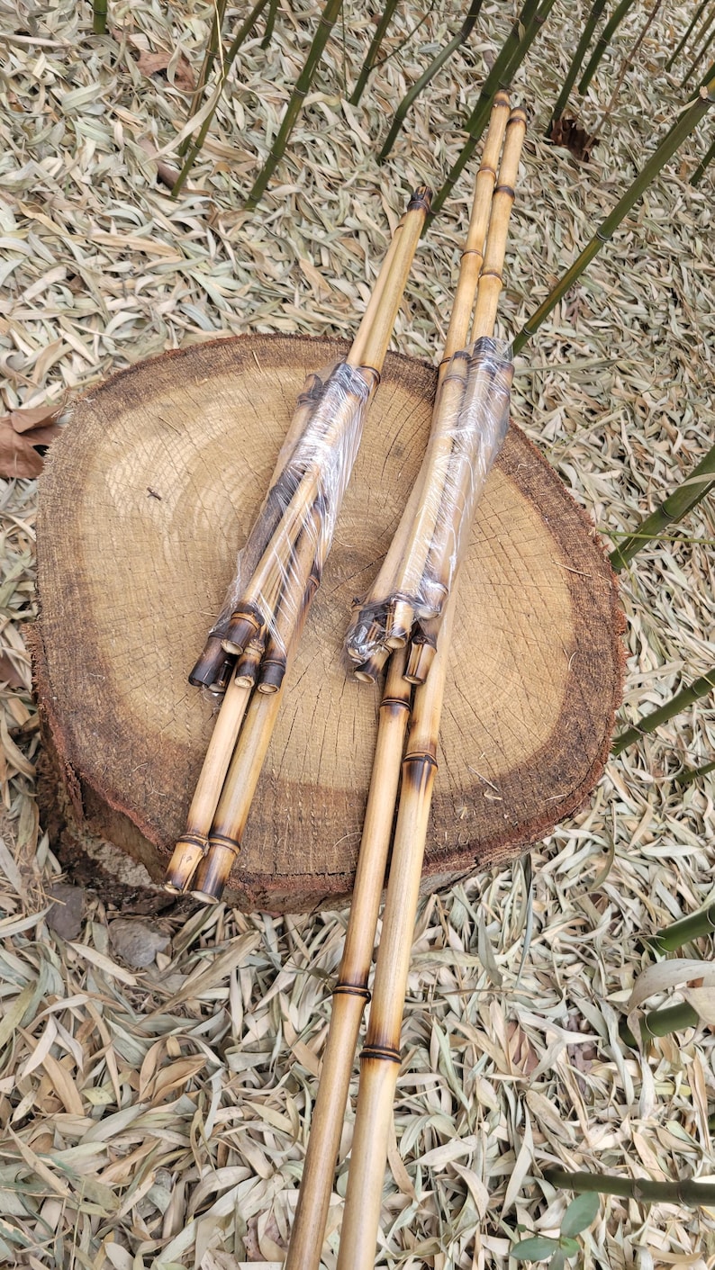 May include: Two bundles of bamboo poles, each wrapped in clear plastic. The bamboo has a natural, light brown color with darker, charred sections at the ends. The poles are resting on a wooden tree stump.