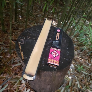May include: A partially burned incense stick resting on a tree stump. The incense stick is black with a white sage and lavender label. The background is a forest of bamboo.