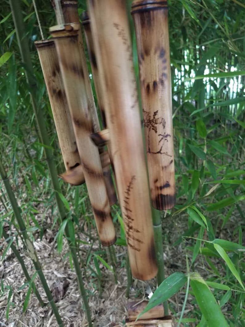 May include: A wind chime constructed from multiple hollow bamboo tubes of varying lengths. The natural tan bamboo features dark, scorched-effect markings and etched illustrations of palm trees and sun motifs on the cylindrical surfaces.