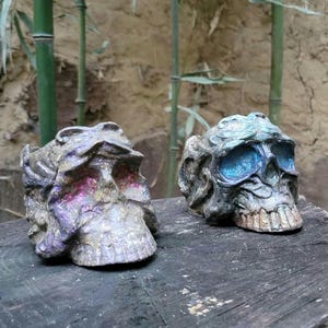 May include: Two decorative skull sculptures, one with a blue and green paint finish and the other with a pink and gold paint finish.