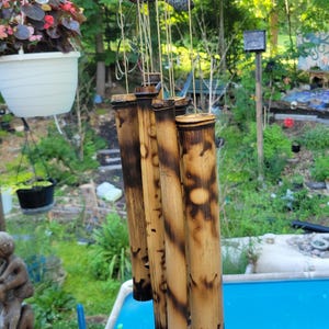 May include: A bamboo wind chime featuring five hollow cylindrical tubes of varying lengths, suspended from a circular wooden top. The bamboo has a natural tan colour with dark, scorched, or burnt-effect markings and patterns along the surface.