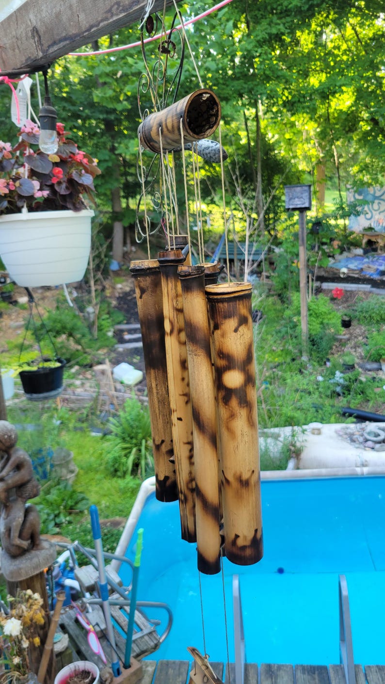 May include: A bamboo wind chime featuring five hollow cylindrical tubes of varying lengths, suspended from a circular wooden top. The bamboo has a natural tan colour with dark, scorched, or burnt-effect markings and patterns along the surface.