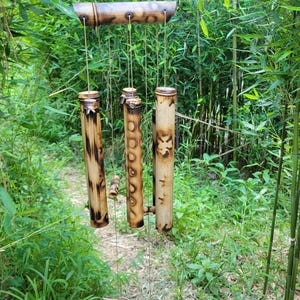 May include: A bamboo wind chime featuring a horizontal top support bar and several vertical hollow bamboo tubes of varying lengths. The natural tan bamboo surfaces are decorated with dark brown scorched patterns and a carved floral design.
