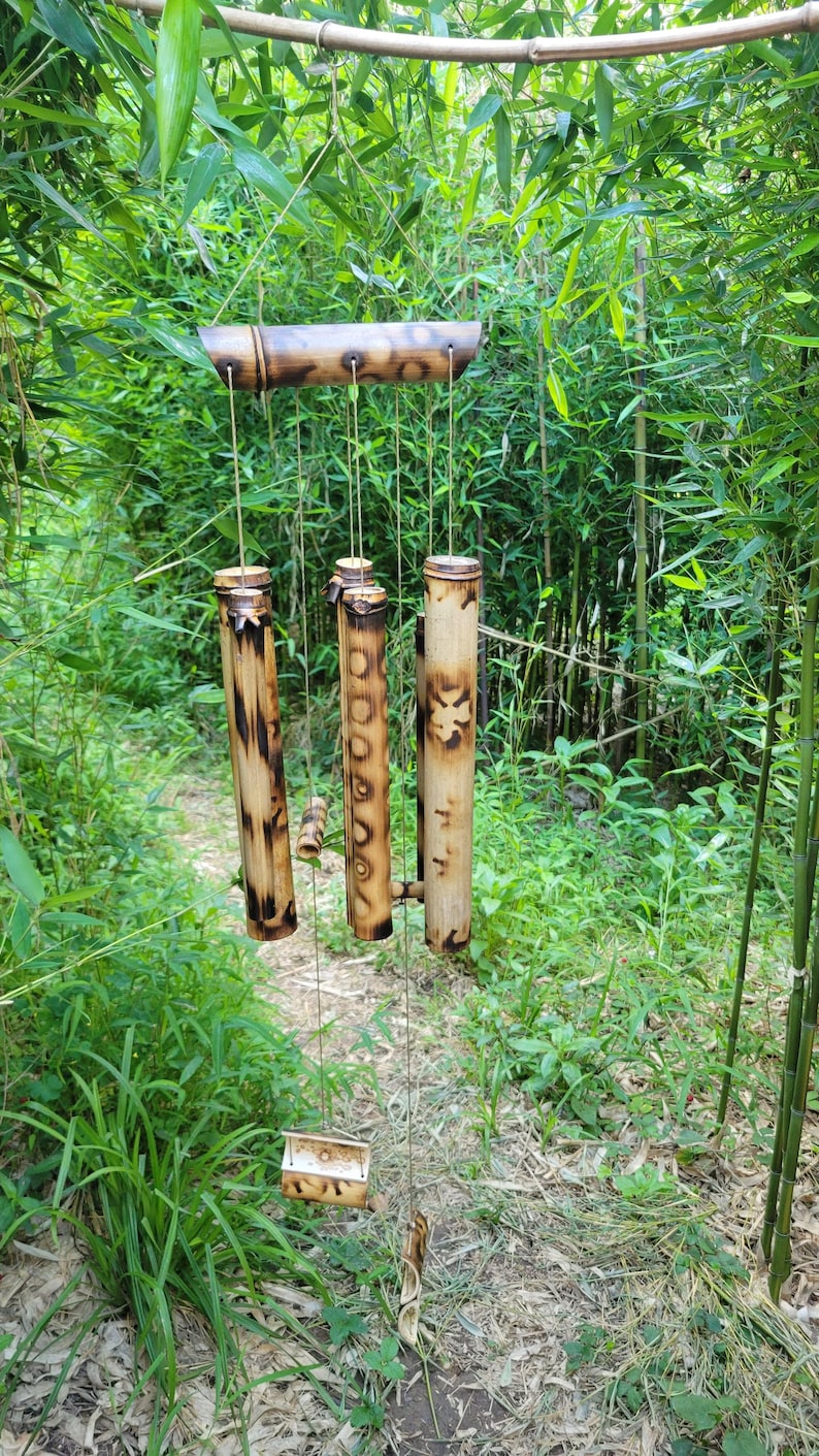 May include: A bamboo wind chime featuring a horizontal top support bar and several vertical hollow bamboo tubes of varying lengths. The natural tan bamboo surfaces are decorated with dark brown scorched patterns and a carved floral design.