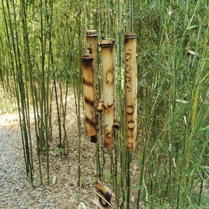 May include: A bamboo wind chime featuring a horizontal top piece and several vertical hollow bamboo tubes of varying lengths. The tubes are decorated with burnt-in circular and geometric patterns, suspended by thin strings from the top support.