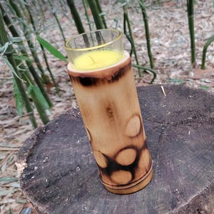 May include: A yellow candle in a glass jar sits inside a bamboo holder with a burnt wood design. The holder is sitting on a wooden surface.