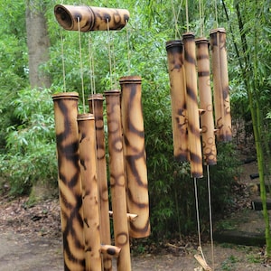 May include: Two bamboo wind chimes hanging from a horizontal branch by red and white plastic clips. Each chime consists of multiple hollow bamboo tubes with dark, scorched-pattern markings, suspended by thin strings from a larger horizontal bamboo piece.