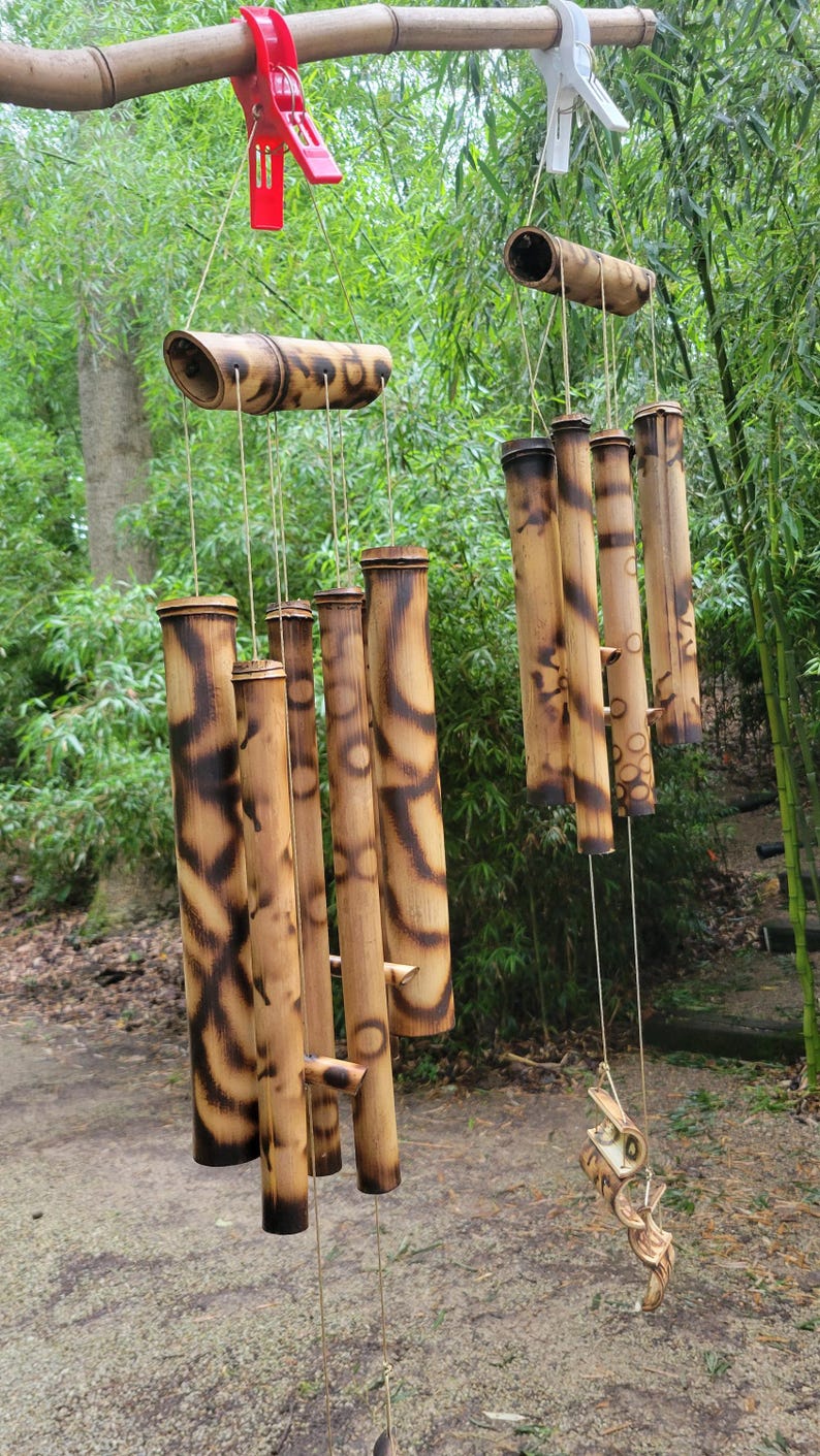 May include: Two bamboo wind chimes hanging from a horizontal branch by red and white plastic clips. Each chime consists of multiple hollow bamboo tubes with dark, scorched-pattern markings, suspended by thin strings from a larger horizontal bamboo piece.