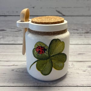 May include: A white, hand-painted jar with a cork lid and a small wooden spoon. The jar features a green four-leaf clover design with a red ladybug. The jar is decorated with twine around the middle.