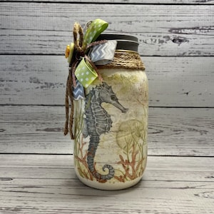 May include: A mason jar decorated with a seahorse and coral design. The jar is tied with a green and white polka dot ribbon and a gray and white chevron ribbon. A small yellow button is attached to the ribbon.