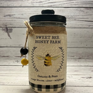 May include: A glass jar with a black lid and a burlap label that reads "Sweet Bee Honey Farm". The label also features a bee illustration and the text "Organic & Pure" and "orange blossom - clover - wildflower". The jar is decorated with a black and white checkered pattern.