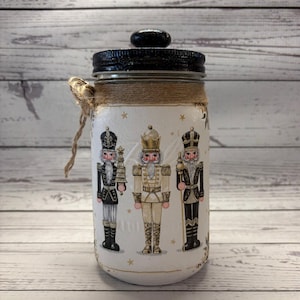 May include: A white mason jar with a black lid and a decorative twine wrap. The jar features three illustrations of nutcracker soldiers in black, gold, and white, with gold star accents. A festive, handcrafted item.