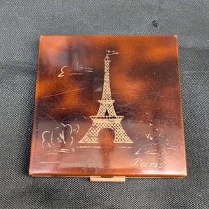 Puede incluir: Un estuche compacto cuadrado con un diseño de carey y un diseño dorado de la Torre Eiffel. La palabra "París" está grabada debajo de la torre. El estuche compacto tiene un pequeño cierre dorado.