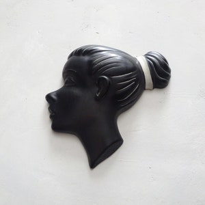 May include: A black wall decor piece in the shape of a woman's profile. The silhouette features a bun hairstyle with a white band. The artwork is mounted on a white textured wall.