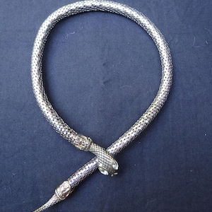 Puede incluir: Un collar de serpiente en tono plateado con un cuerpo flexible tipo malla y un patrón de escamas texturizado. Los dos extremos del collar tienen forma de cabeza de serpiente, con una de las cabezas adornada con pequeñas piedras claras.